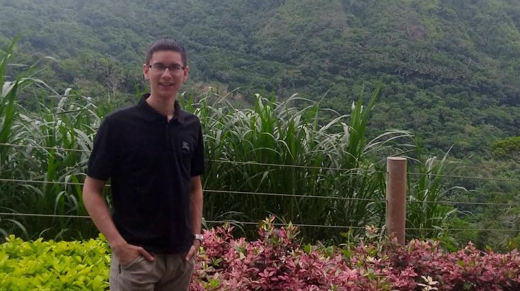 Alum Raphael Roxas in a polo and shorts standing in front of a green field and hills.